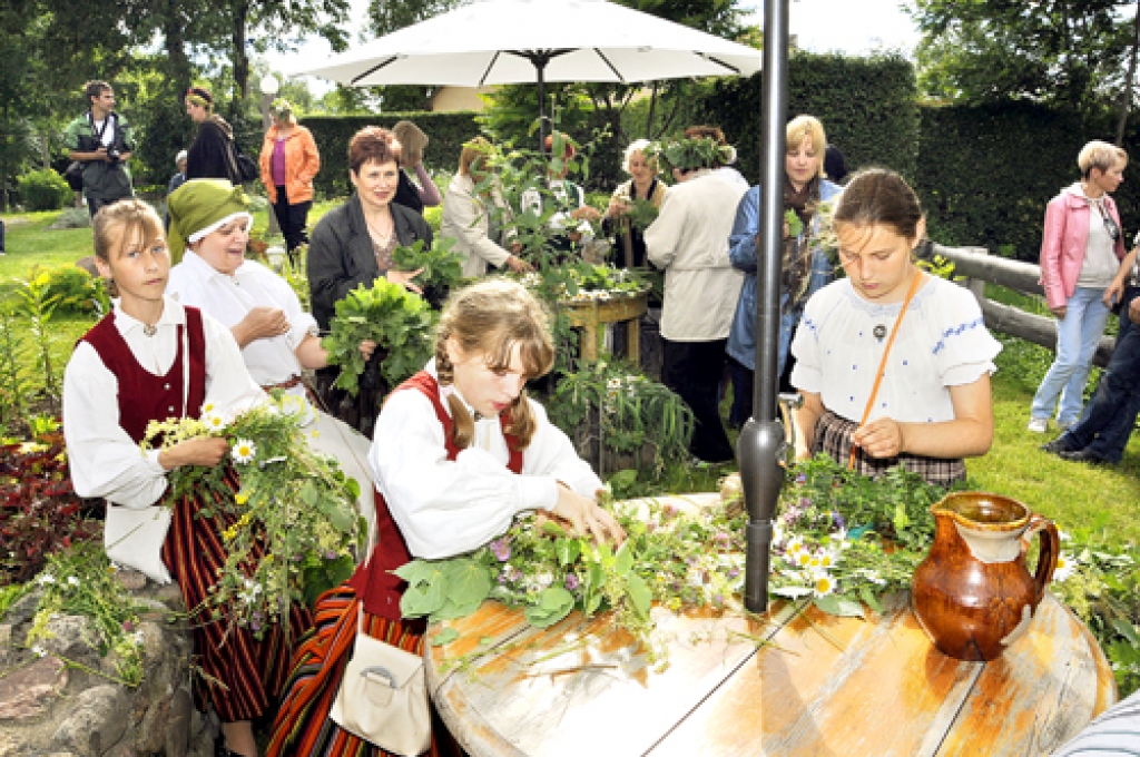 Baltās mājas saimnieki Ikerti un Silmaču saime māca līgotājiem svētku aktivitātes, arī Jāņu vainagu pīšanu.
