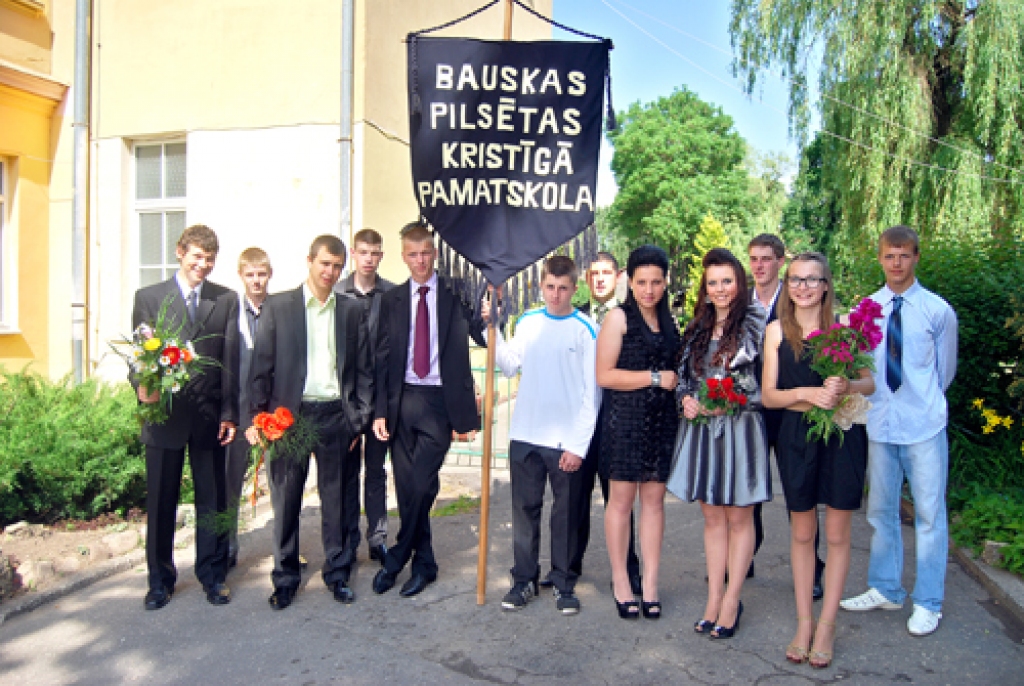 Brīdi pirms apliecību saņemšanas pie Kristīgās pamatskolas piestājuši tās pēdējie absolventi. Viņi nes mācību iestādes karogu.  