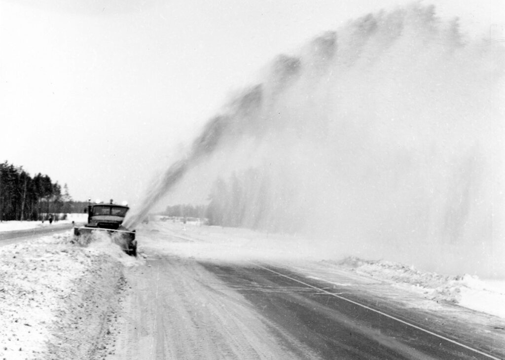 Sniega novākšana Pierīgas maģistrālēs ar sniega pūtēju, 1970. gadi. Smagā automašīna ar lāpstu un rotoru sniega valni aizpūš tālu pāri vairāku joslu ceļam. Foto no Latvijas Ceļu muzeja.